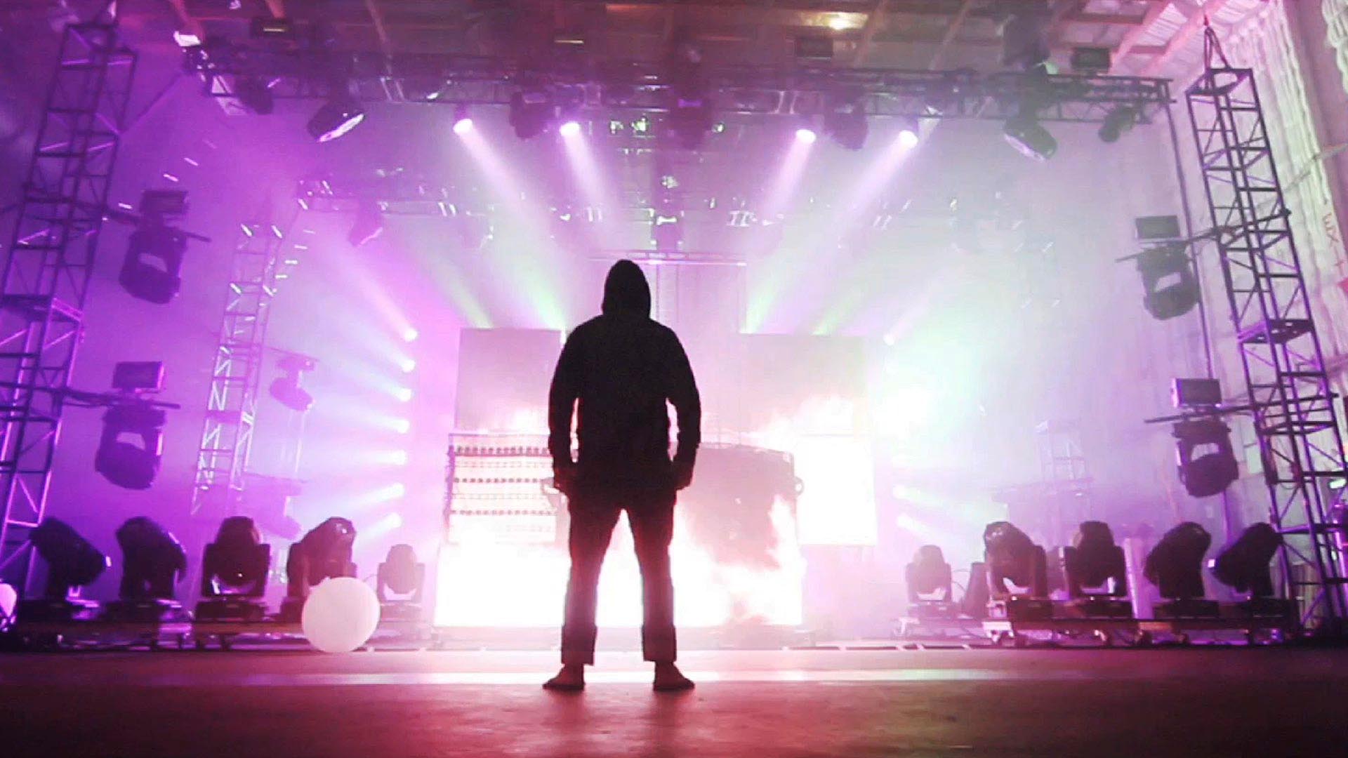 Silhouette of a person in a hoodie standing before a brightly lit stage with pink and purple lights.