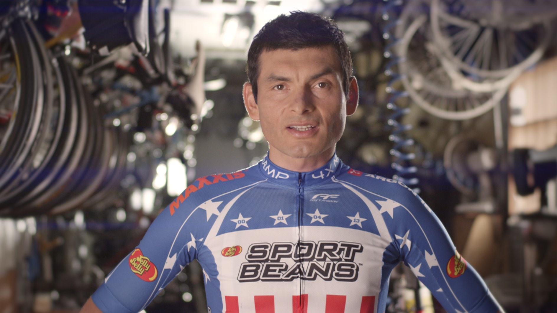 Cyclist in a star-spangled jersey stands in a bike workshop, with tools and wheels in the background.