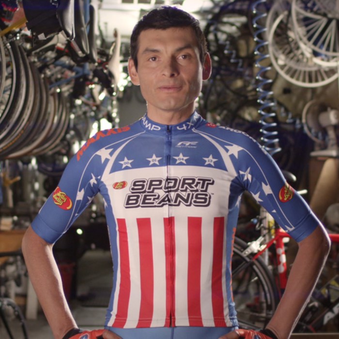 Cyclist in a star-spangled jersey stands confidently in a cluttered bicycle workshop.