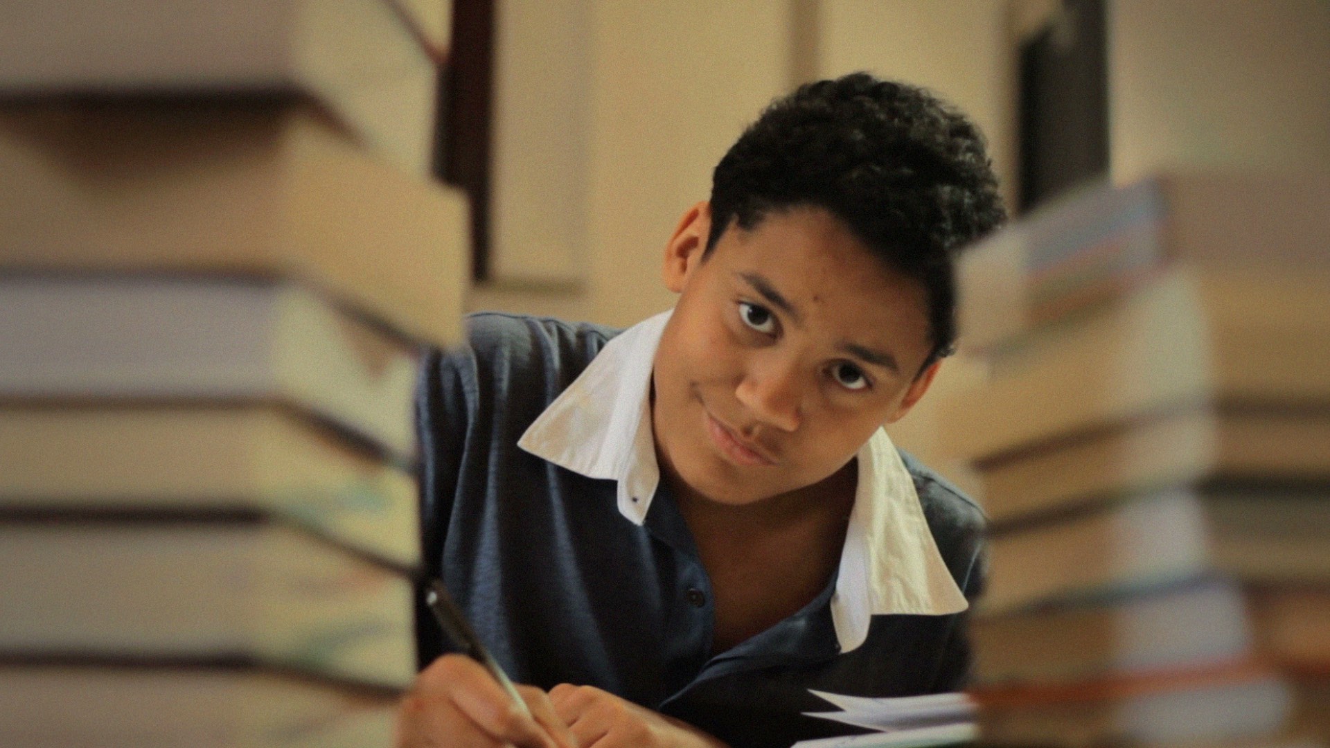 Person writing at a desk, surrounded by tall stacks of books, peering curiously between them.