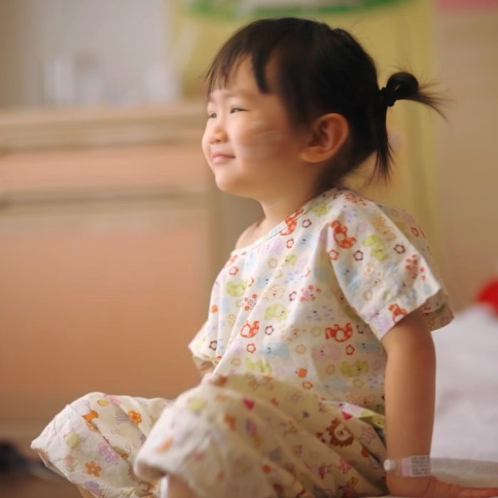 Smiling toddler with ponytail sitting, wearing floral hospital gown in a room.