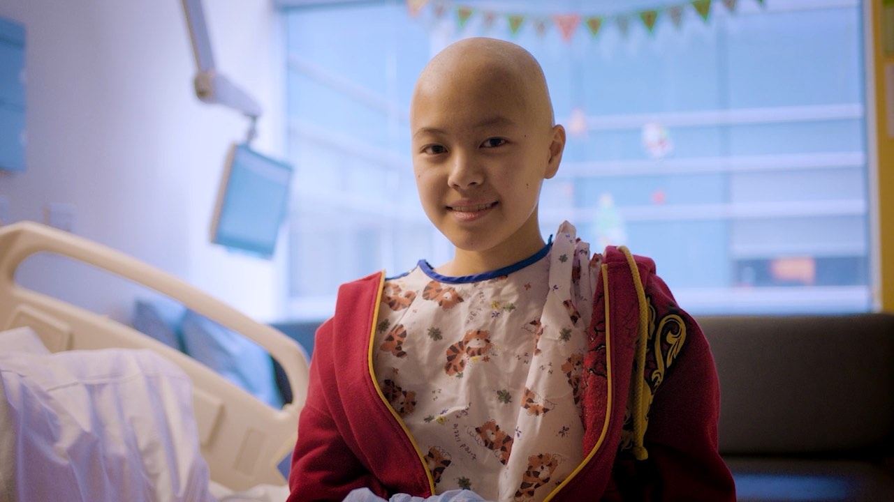 Child in hospital room, wearing a red hoodie and floral gown, smiling while sitting on a bed.