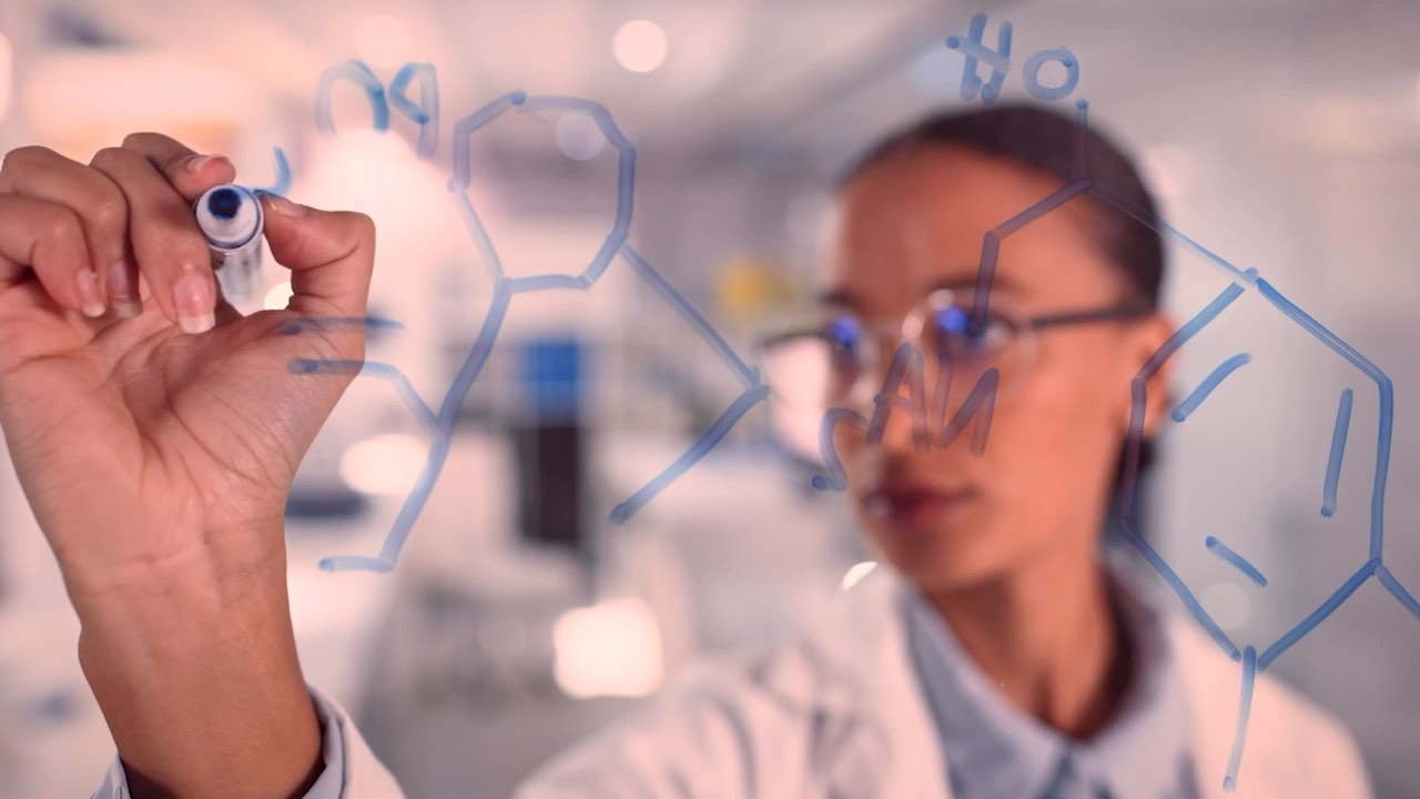 Scientist writing chemical formulas on transparent board in a laboratory setting.