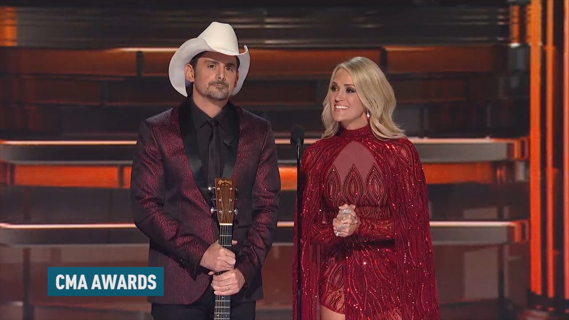 A man and woman in evening attire stand on stage at the CMA Awards. The man holds a guitar.