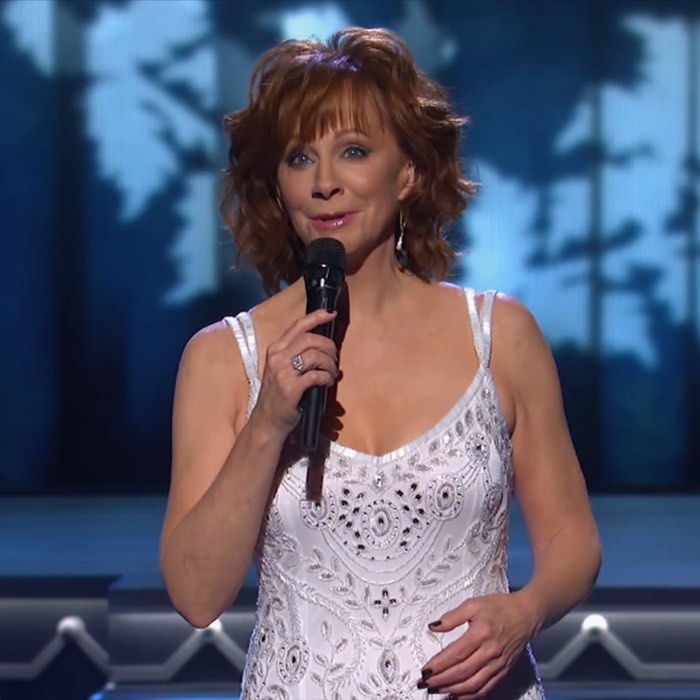 A woman in a white dress holds a microphone on stage with a forest backdrop.