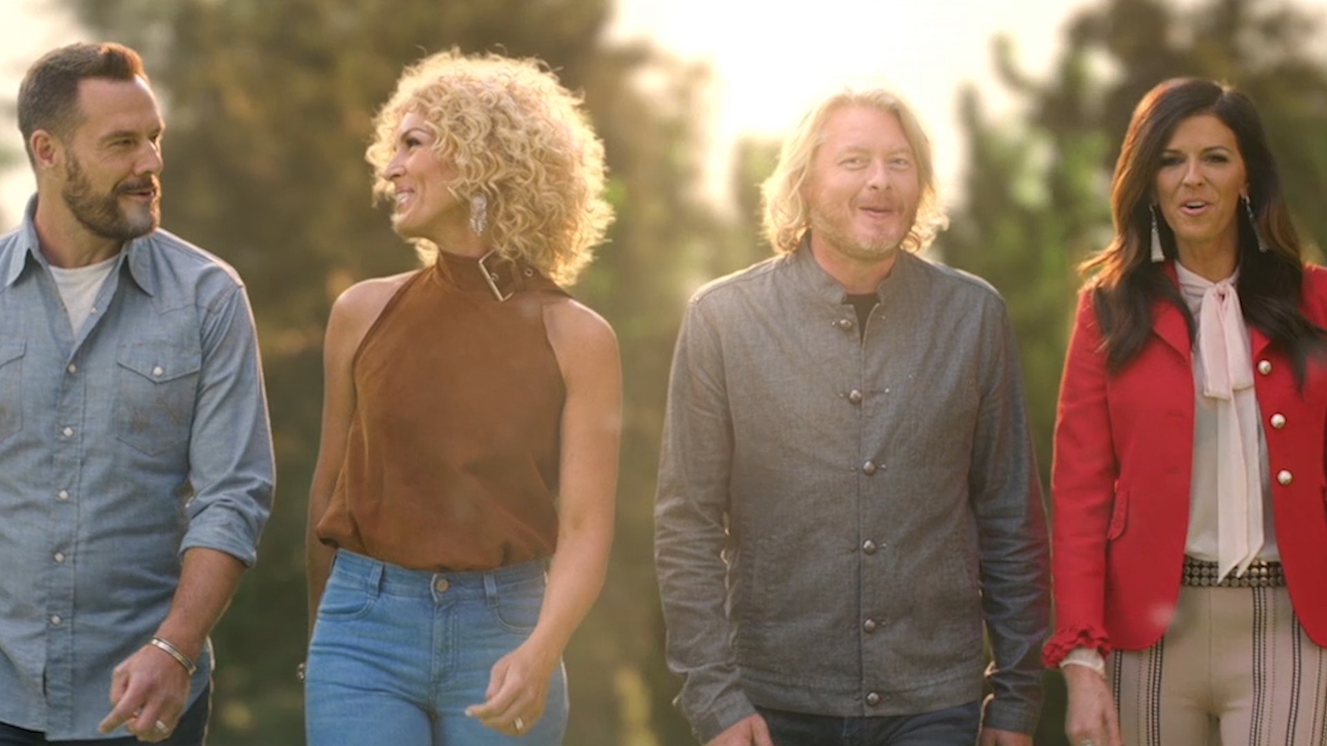 Four people walking outdoors, smiling, with trees in the background and sunlight filtering through.