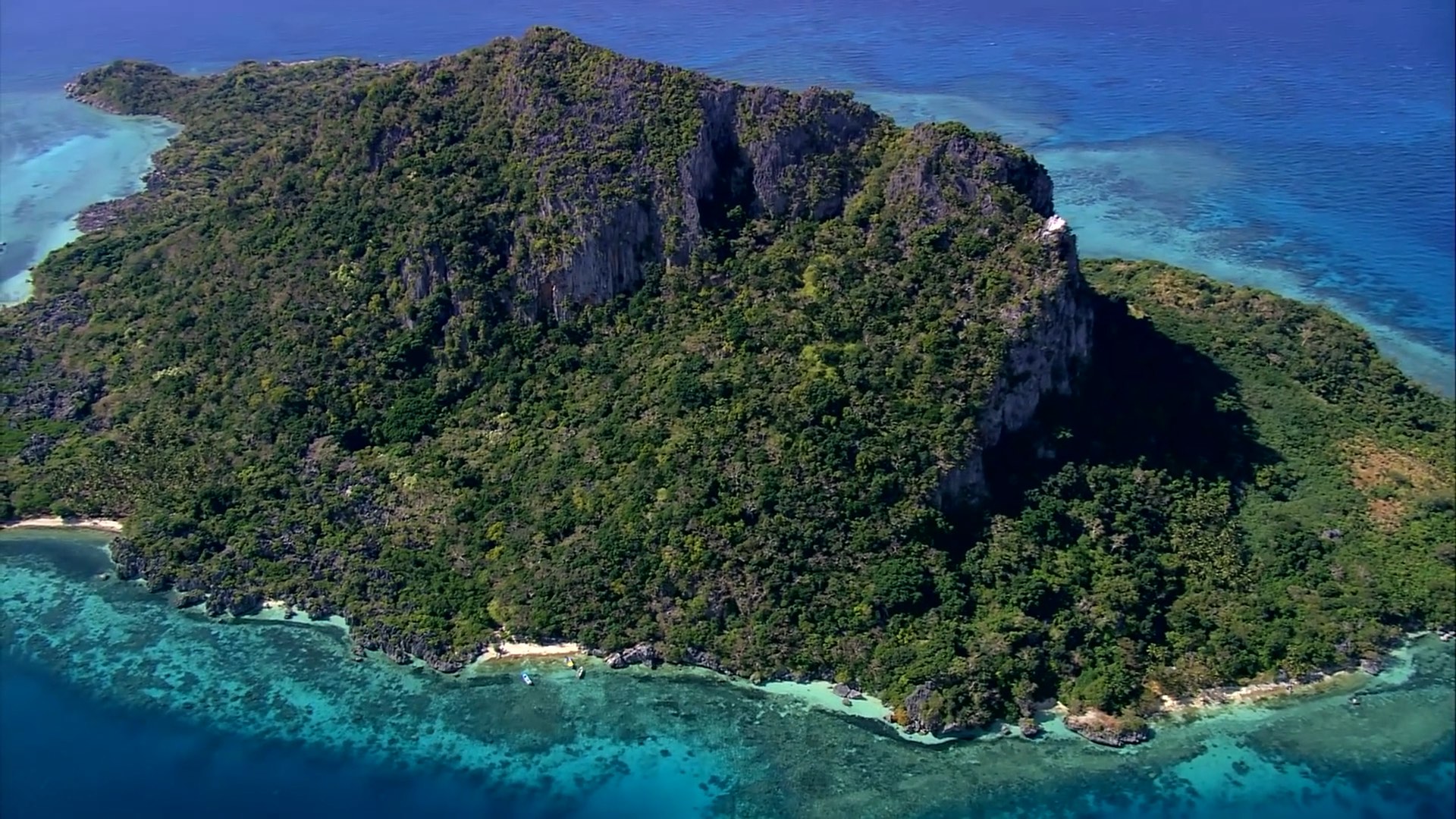 A lush, green island with steep cliffs surrounded by turquoise ocean waters under a clear blue sky.