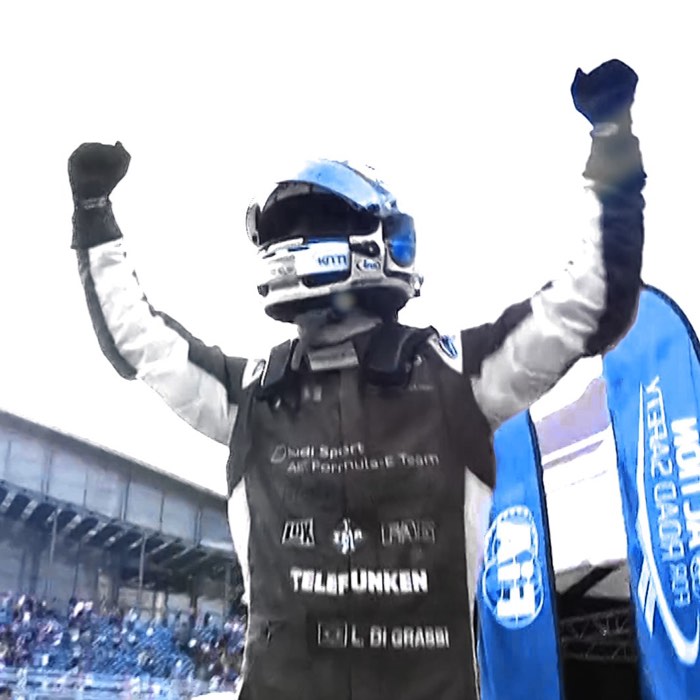 Race car driver celebrating with arms raised, wearing a racing suit and helmet, blue banner in background.