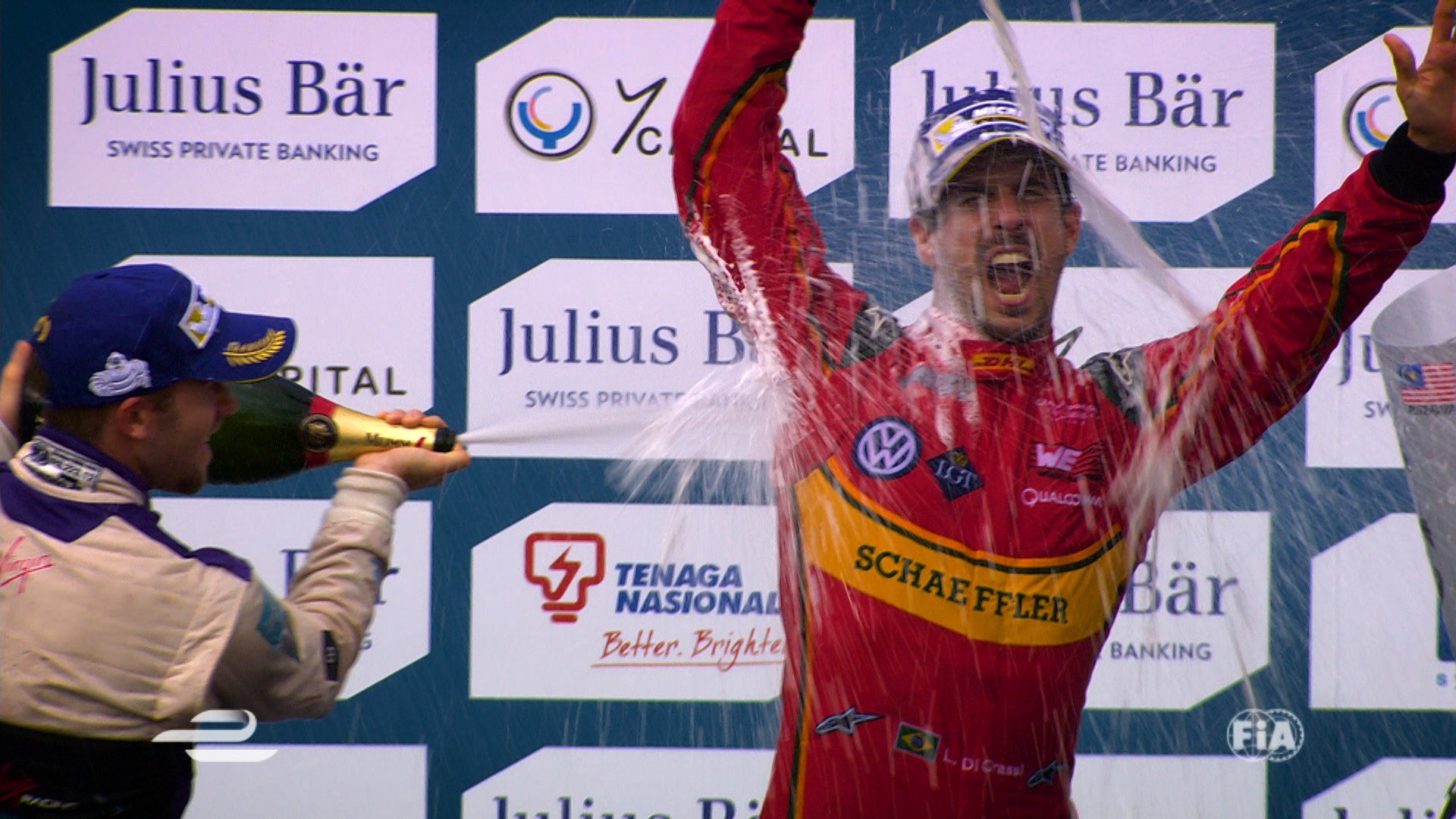 Race car driver celebrating on podium with champagne, arms raised. Another driver sprays him with champagne.