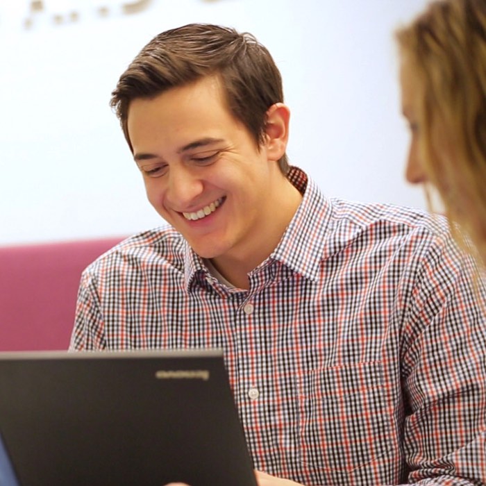 A person smiling at a laptop, sitting next to someone else, in a casual setting.