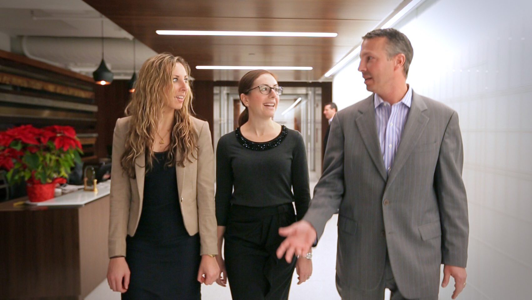 Three professionals walk and talk in an office corridor, with holiday decorations to the left.