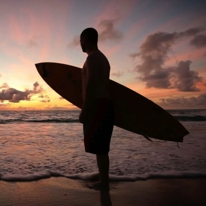 Surfer silhouetted against a colorful sunset, standing on a sandy beach with a surfboard.