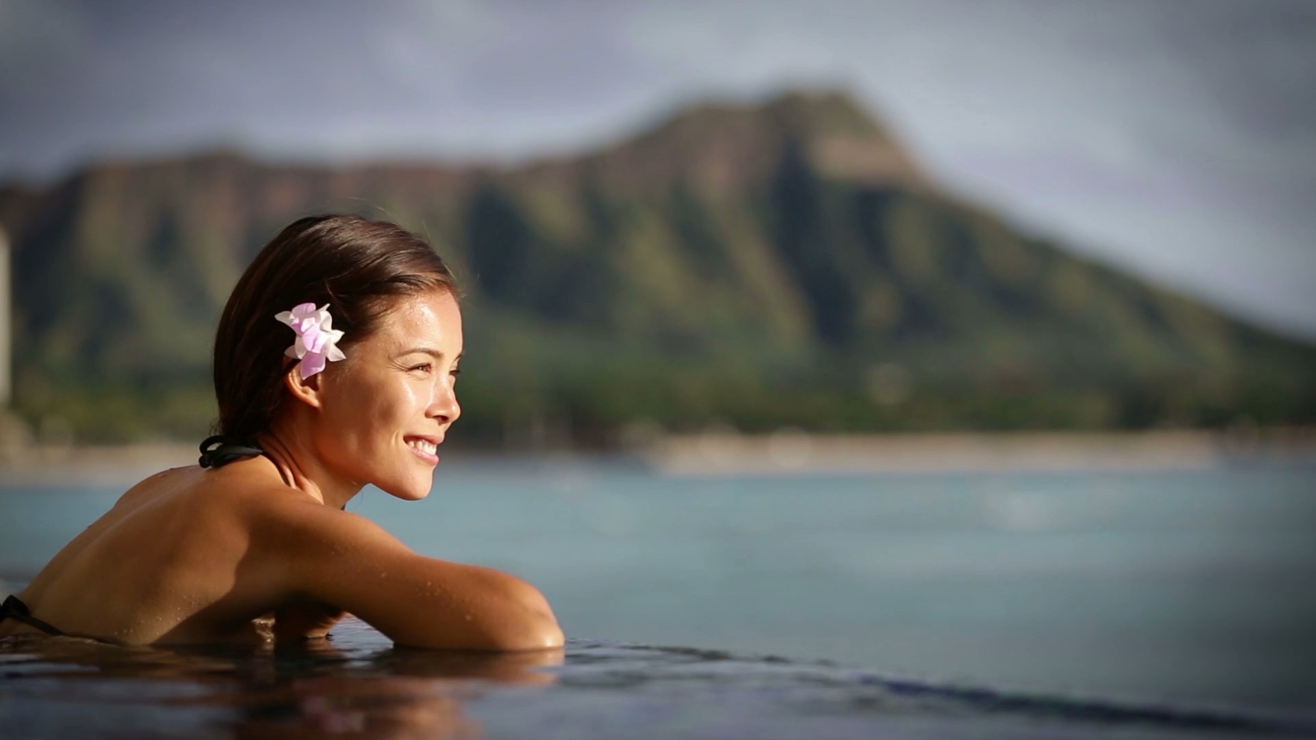Smiling woman in a pool with a flower in her hair, mountain in the background.