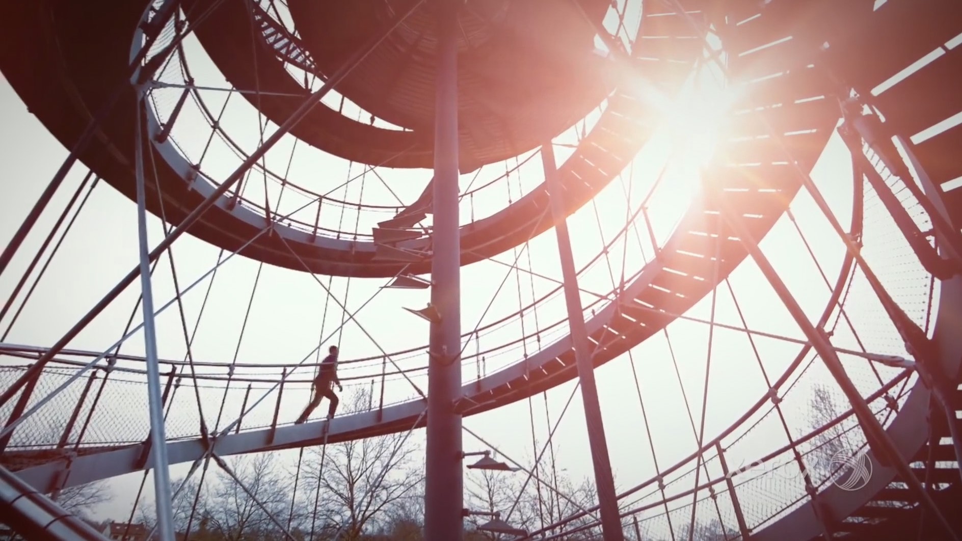 Person walks on a spiral staircase structure in sunlight, with trees visible in the background.