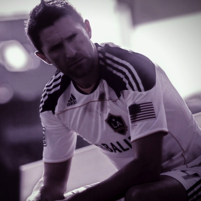 Soccer player in white LA Galaxy uniform sits pensively on the bench in a moody, black-and-white photo.