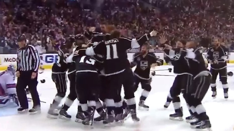 Hockey team celebrating on ice in black uniforms as referee watches, with crowd in the background.