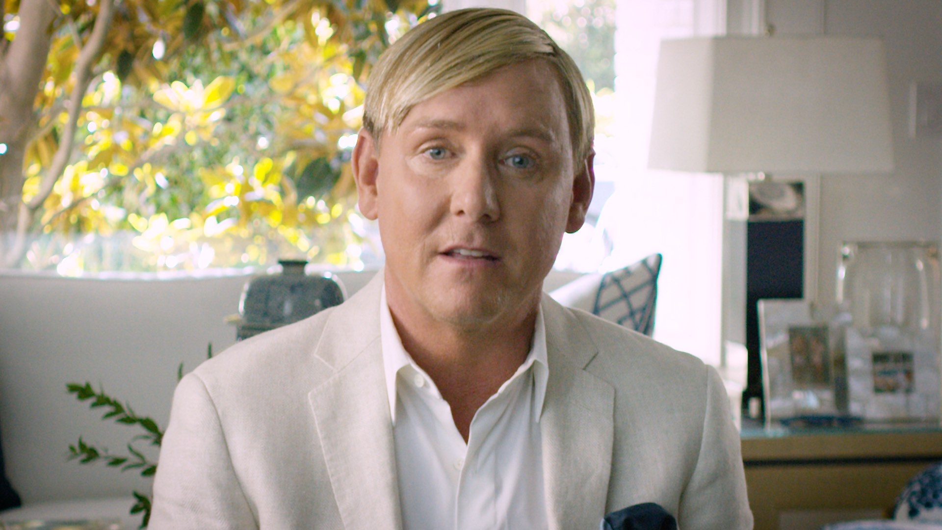 Person in a beige jacket and white shirt sitting indoors with plants and a lamp in the background.