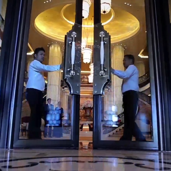Two doormen in white shirts open large glass doors at an elegant hotel entrance with ornate lighting.