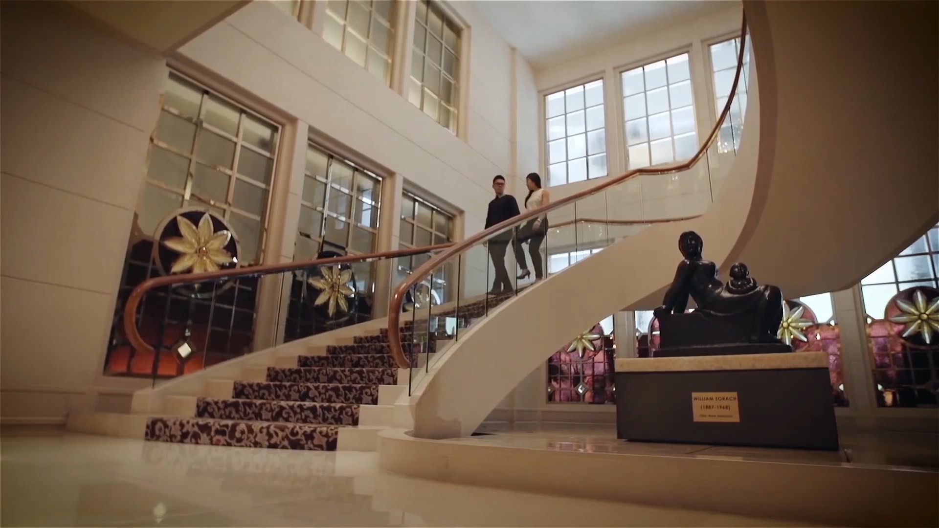 Two people stand on a grand staircase with ornate railing, next to large windows and a modern sculpture.