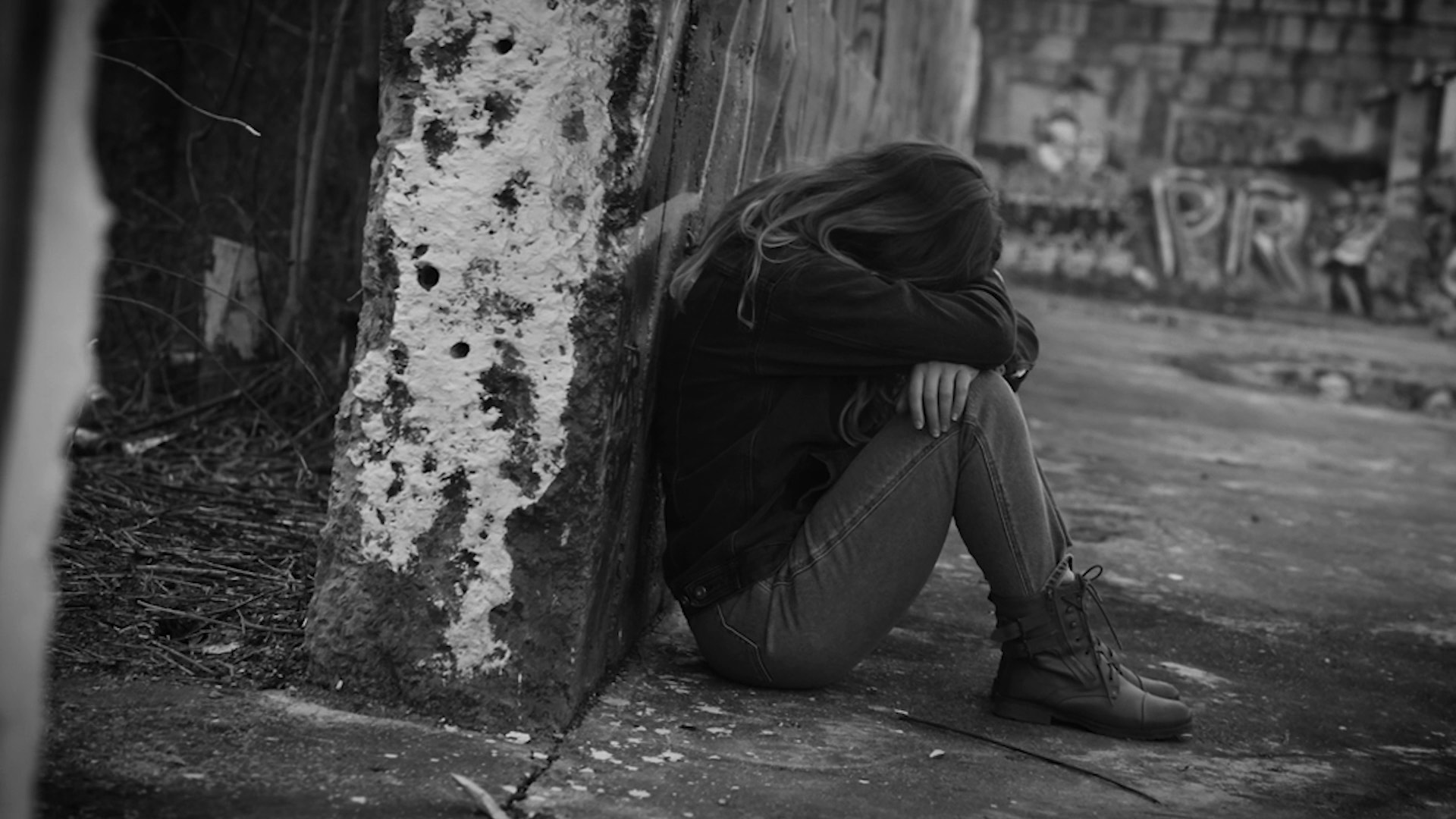Person sitting on the ground against a wall, head down, in a desolate, graffiti-covered urban setting.