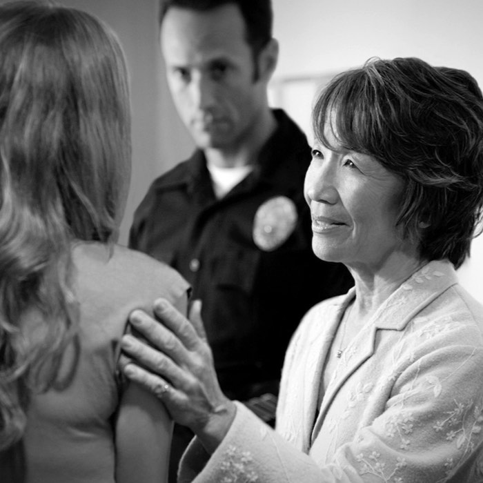 A smiling woman comforts another woman while a police officer stands in the background. Black and white image.
