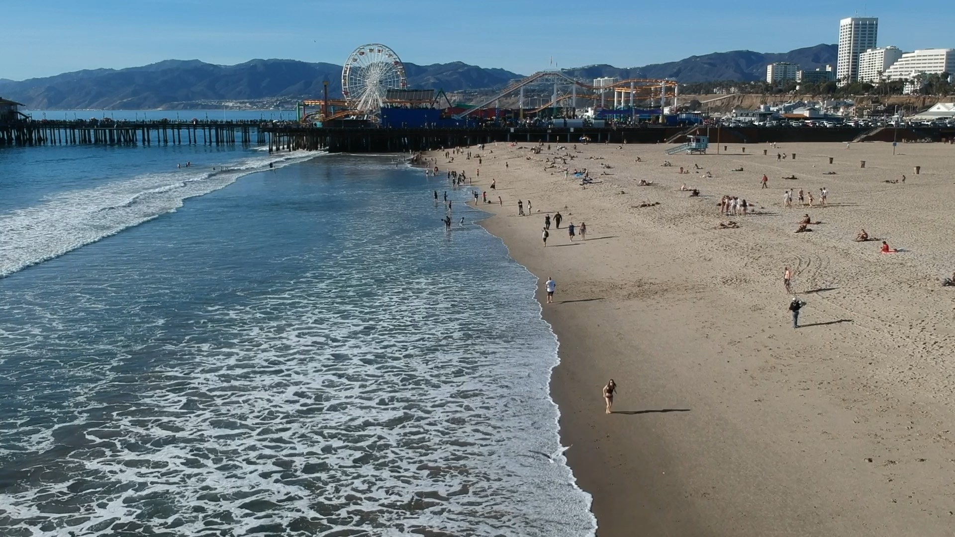 A beach with people, a pier, and a ferris wheel in the distance under a clear blue sky.