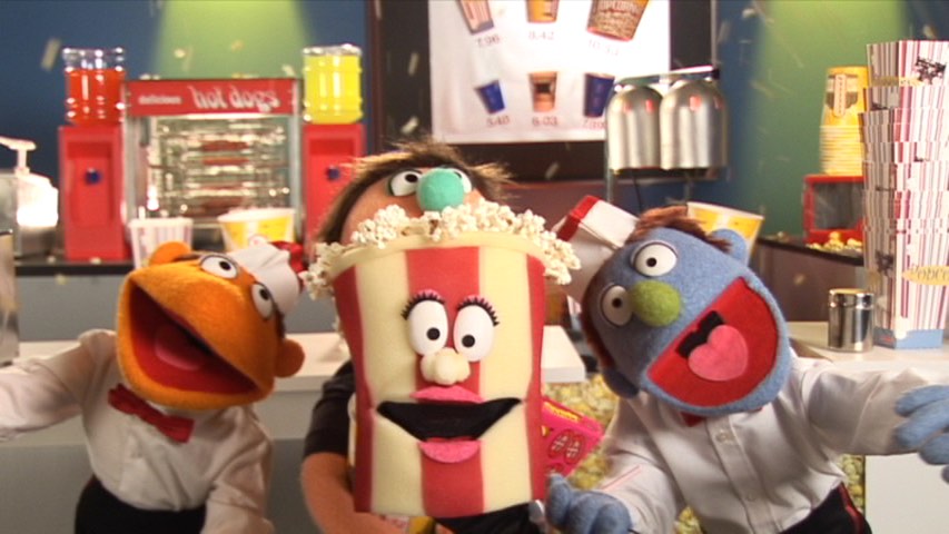 Colorful puppets in a retro food stand, one shaped like a popcorn bucket with makeup and a smiling expression.