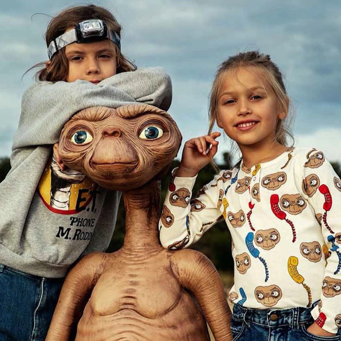 Two children are posing outdoors with an E.T. figure. They're both smiling, and the sky is cloudy.
