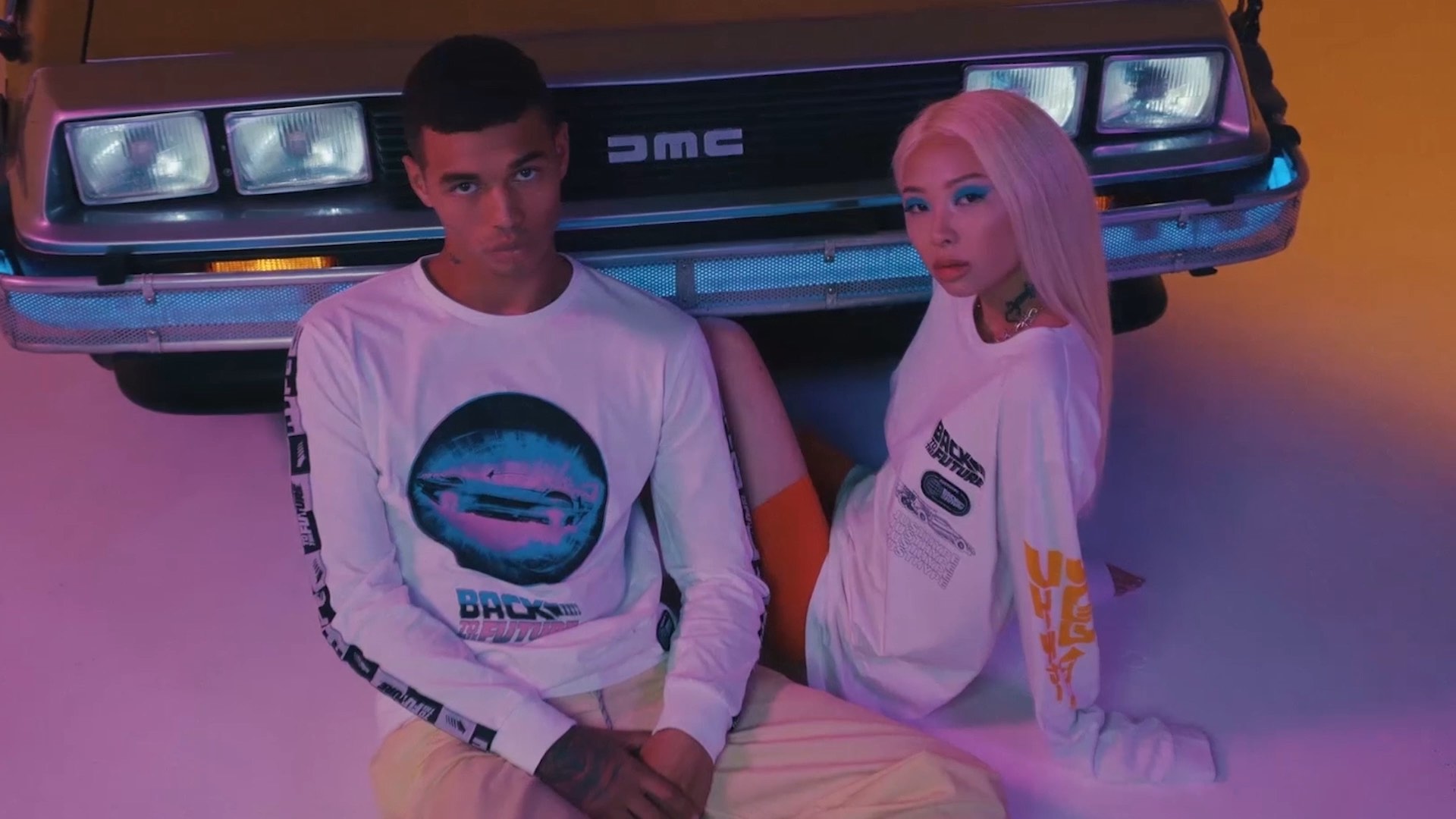 Two people in graphic shirts sit on the floor in front of a DMC car, with colorful lighting.
