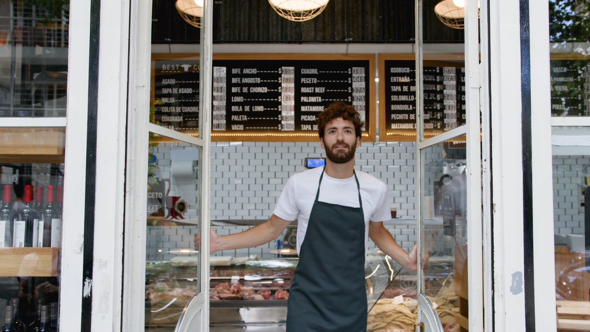 Man in an apron stands at a butcher shop entrance with a menu board above and meat displayed inside.