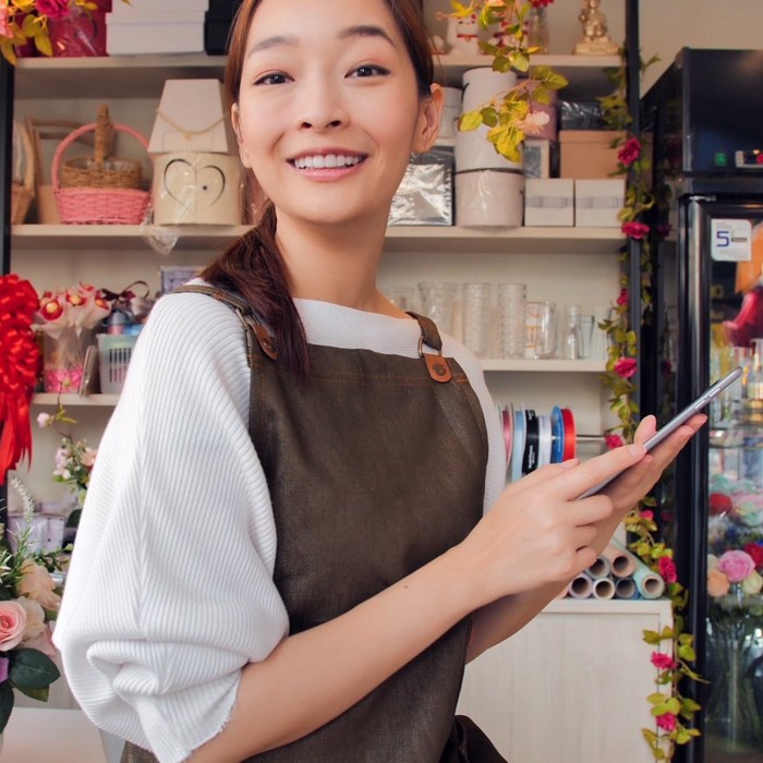 Smiling person in an apron stands in a floral shop, holding a smartphone.