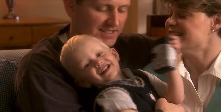 Smiling child sitting on a man's lap with a woman nearby, all appearing happy and relaxed indoors.