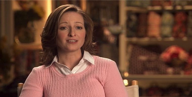 Woman in a pink sweater sits and talks in a cozy room with shelves in the background.